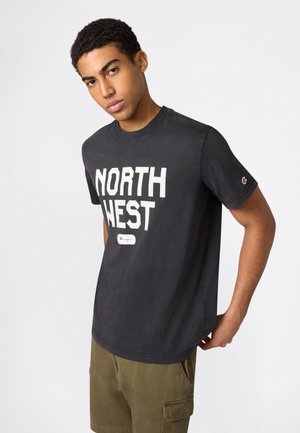 Młody mężczyzna ubrany w czarną koszulkę z napisem „NORTH WEST” i oliwkowe szorty typu cargo, stojący z pochyloną głową i jedną ręką w kieszeni.