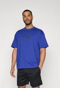 Mėlynas medvilninis marškinėliai su juodu Nike logotipu ant krūtinės. Trumpomis rankovėmis, laisvas kirpimas ir apvalus iškirpimas. Dėvimi su juodomis šortais.