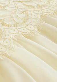 Kollane pitskangas, millel on keerukas lillemuster ja frillides servad, on kihiline sujuva siidise materjali kohal, rõhutades tekstuuri ja õrna disaini.