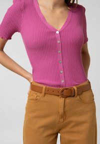 Žebrovaný top s krátkým rukávem v živé růžové barvě s výstřihem do V a knoflíkovým zapínáním, kombinovaný s béžovými kalhotami se páskem vyrobenými z měkké tkaniny.