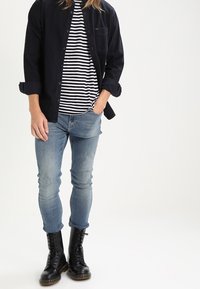 Camicia blu navy con bottoni sopra una maglietta a righe bianche e nere, jeans azzurri chiari e stivali neri in pelle con lacci. Tessuto morbido, vestibilità casual.