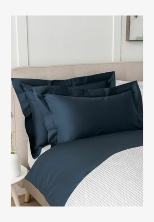 Beiges gepolstertes Bett mit dunkelblauen Kissenbezügen und Bettbezug neben einem weißen Nachttisch, auf dem eine weiße Vase mit einer grünen Pflanze steht.