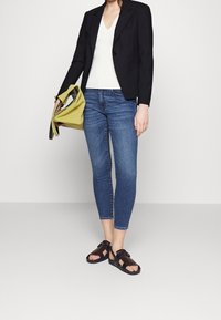 Černý blazer, bílý top s V-výstřihem, modré kotníkové džíny a hnědé sandály. Outfit doplňuje zelená taška s texturovaným povrchem.