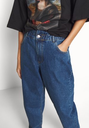 Person wearing loose blue jeans with elastic waistband and black t-shirt featuring Mona Lisa print, hands in pockets.