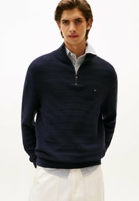Maglione navy a mezza zip in maglia rib ribattuta, con una camicia a righe sotto. Dettaglio logo discreto sul petto. Tessuto morbido e design aderente.