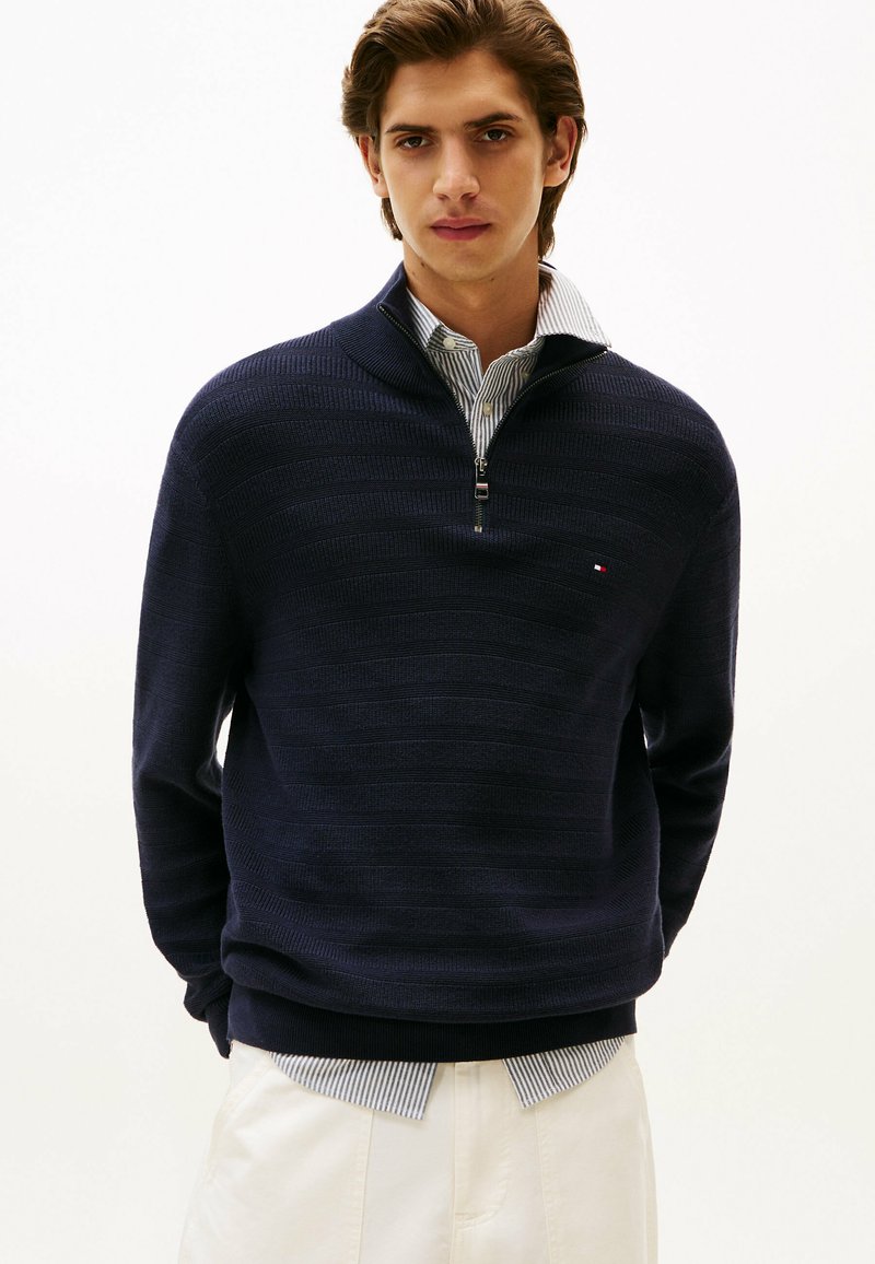 Maglione navy a mezza zip in maglia rib ribattuta, con una camicia a righe sotto. Dettaglio logo discreto sul petto. Tessuto morbido e design aderente.