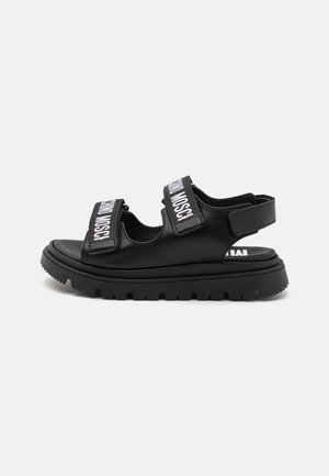 Sandalias negras con dos correas anchas que presentan una marca blanca. Suela de goma gruesa con patrón de textura, diseño de talón abierto.