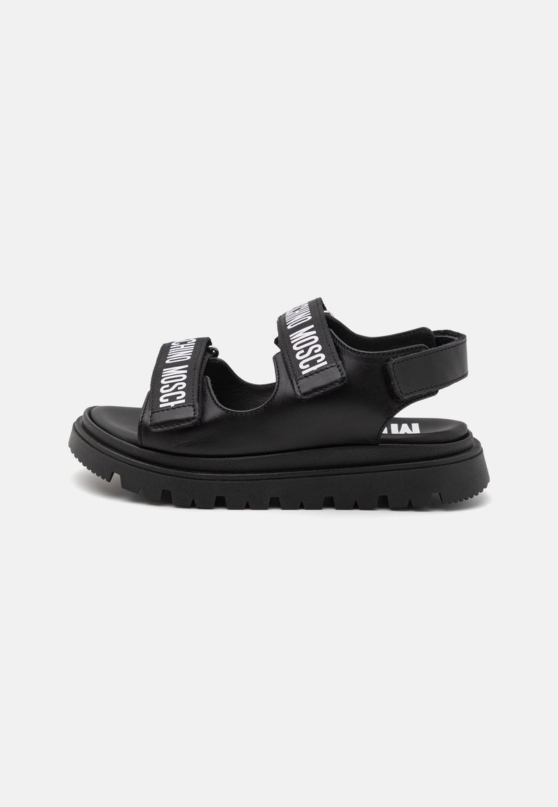 Sandalias negras con dos correas anchas que presentan una marca blanca. Suela de goma gruesa con patrón de textura, diseño de talón abierto.