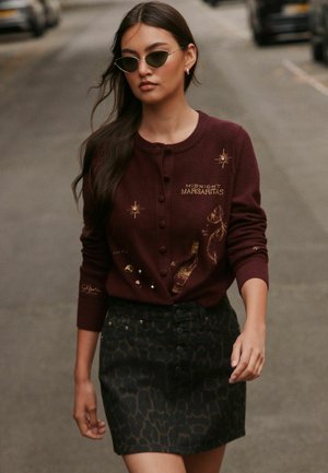 Mujer con gafas de sol estilo cat-eye, cárdigan burdeos con texto "Midnight Margaritas" y falda oscura con estampado caminando por la calle.
