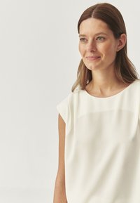 Blusa ligera color crema con cuello redondo, mangas cortas y textura suave. Presenta un brillo sutil y un diseño minimalista.