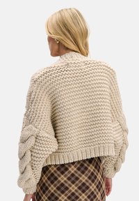 Kremowy dziany sweter o grubym splocie, szerokie rękawy z plecionym wzorem oraz krótki krój. Gładki tył z wysokim kołnierzem.