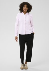 Chemise à rayures rose clair avec boutons et col, associée à un pantalon noir et des baskets beiges. La chemise est ajustée et à manches longues.
