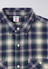 Camicia di flanella a quadri blu navy e verde con accenti bianchi, dotata di colletto con bottoni, due taschini frontali e un'etichetta di marca all'interno del colletto.