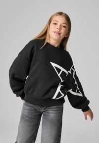 Svart sweatshirts med en stor vit stjärngrafik, ribbade muddar och nederkant, i kombination med grå jeans. Ytan verkar mjuk och texturerad.