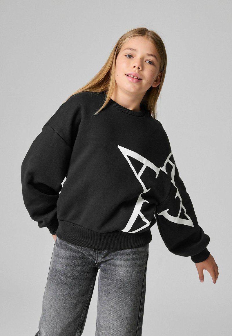 Svart sweatshirts med en stor vit stjärngrafik, ribbade muddar och nederkant, i kombination med grå jeans. Ytan verkar mjuk och texturerad.
