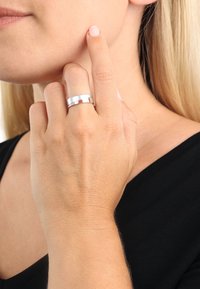 Anello d'argento con una finitura lucida, caratterizzato da un design piatto, indossato sul dito anulare, accanto a una mano che tocca la guancia.