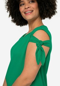 Groen T-shirt met korte mouwen en een geknoopte detail op de schouder. Zachte stof, ontspannen pasvorm en ronde hals zorgen voor comfort en stijl.