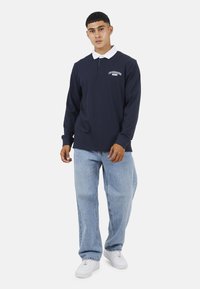 Polo a maniche lunghe blu navy con colletto bianco e logo, abbinato a jeans azzurri e sneakers bianche. Materiale in cotone; vestibilità rilassata.