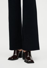 Γυαλιστερά σκούρα καφέ δερμάτινα mule με τακούνι, φορεμένα με μαύρο παντελόνι φαρδιάς γραμμής σε ανοιχτό φόντο.