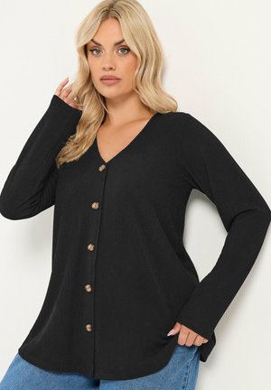 Blond kvinde iført en sort langærmet knapbluse med blå jeans, poserende mod en ensartet lys baggrund.