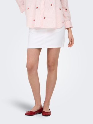 Vrouw met een witte minirok, een roze blouse met lange mouwen met kleine kersenprints en rode ballerina's, staand tegen een grijze achtergrond.