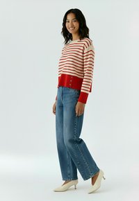Femme souriante portant un pull rayé rouge et blanc, un jean large bleu, et des chaussures crème à petit talon, sur un fond uni.