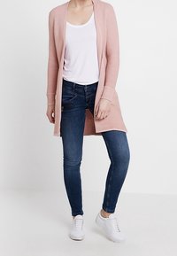 Cardigan a costine rosa chiaro con apertura frontale, abbinato a una maglietta bianca e jeans blu scuro. Sneakers bianche completano l'outfit.
