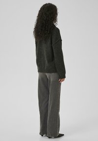 Woman with curly dark hair wearing a dark gray sweater, loose gray jeans, and black pointed heels, standing facing away on a plain background.