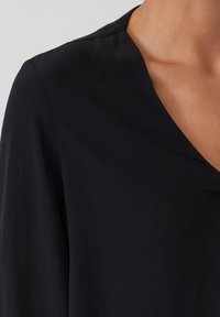 Schwarzes Oberteil mit V-Ausschnitt, weichem Material und leicht drapierten Ärmeln, das eine klare und moderne Silhouette präsentiert.