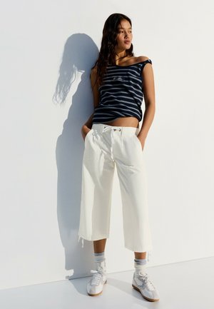 Vrouw staat tegen een witte muur en draagt een mouwloos gestreept topje, witte wijde cropped broek, grijze sokken en witte sneakers.