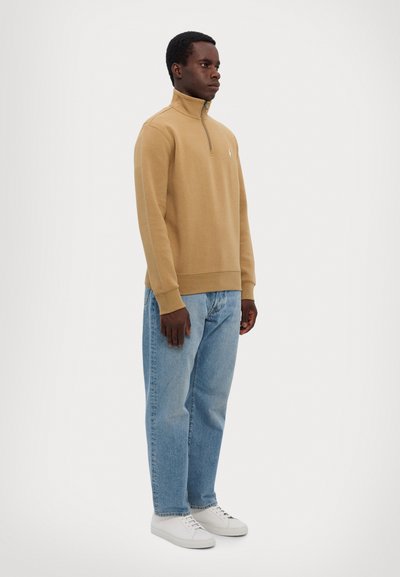 Tan kvart-zip sweatshirt lavet af blødt stof, parret med lyseblå lige ben jeans og hvide sneakers, der udviser en afslappet pasform.
