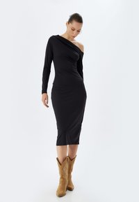 Robe noire à manches longues avec un col asymétrique, silhouette ajustée et ourlet au niveau des genoux, associée à des bottes de cow-boy beige ornées de broderies.