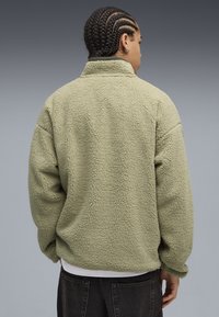 Giacca in fleece di colore verde chiaro con superficie morbida e texture. Presenta un colletto alto e una vestibilità oversize con spalle cadenti.