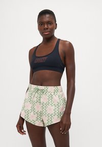 Atletski crop top u tamnoplavoj boji s crvenim logotipom "ROXY", u kombinaciji sa svijetlozelenim šortsevima koji imaju ružičaste cvjetne i valovite uzorke. Meki materijal, elastična sredina.