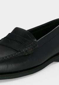 Μαύρο δερμάτινο penny loafer με ραφές moc toe και επίπεδη μαύρη σόλα, εμφανίζεται σε κοντινό πλάνο σε ανοιχτό φόντο.