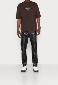 Brązowy, oversize T-shirt z graficznym nadrukiem na piersi, połączony z błyszczącymi, czarnymi spodniami z imitacji skóry i białymi butami na sznurówki.