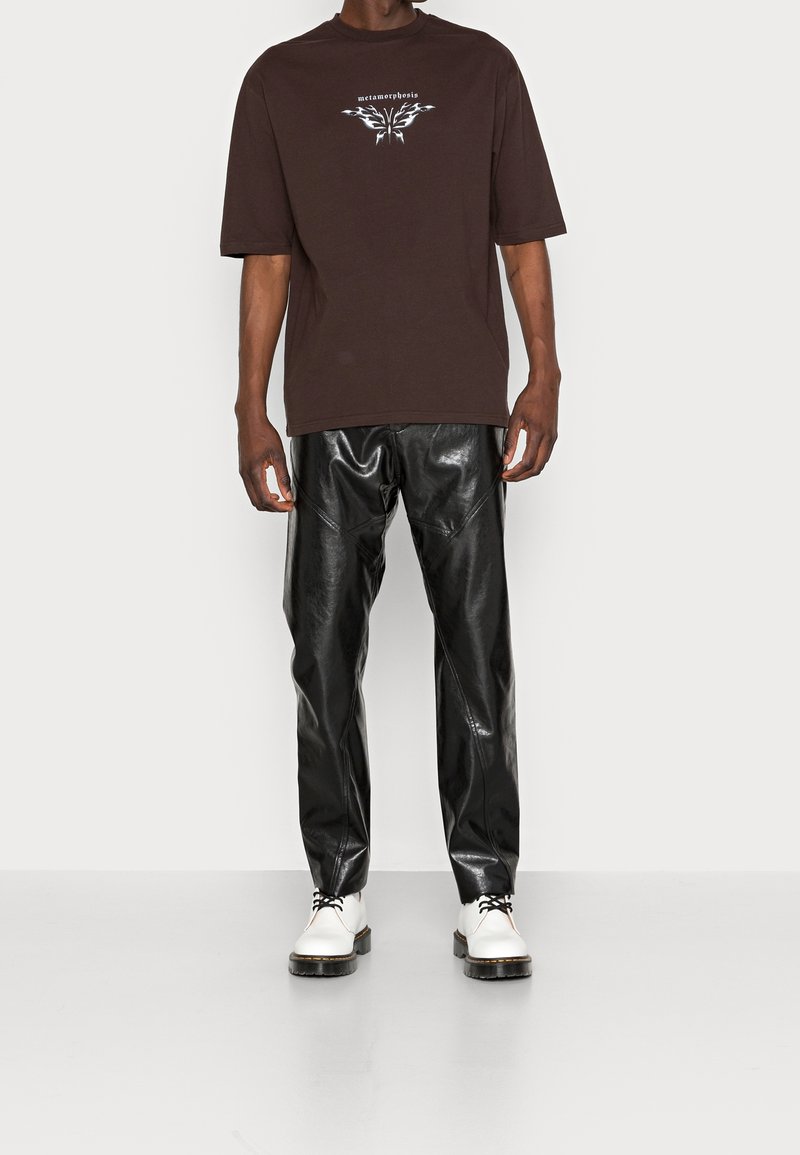 Brązowy, oversize T-shirt z graficznym nadrukiem na piersi, połączony z błyszczącymi, czarnymi spodniami z imitacji skóry i białymi butami na sznurówki.