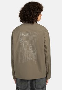Light brown jacket with a grid-patterned white graphic on the back. Features a smooth texture and long sleeves, designed for casual wear.