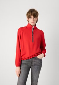 Rode lichtgewicht pullover met een halve rits, elastische onderkant en lange mouwen. Voorzien van zwart-witte merkletters op de rits.