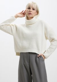 Pull à col roulé en tricot côtelé couleur crème, de coupe courte et décontractée, associé à un pantalon gris taille haute.
