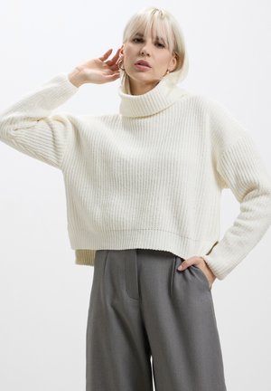 Maglione a collo alto a costine di colore crema, con un design cropped e una vestibilità rilassata, abbinato a pantaloni grigi a vita alta.