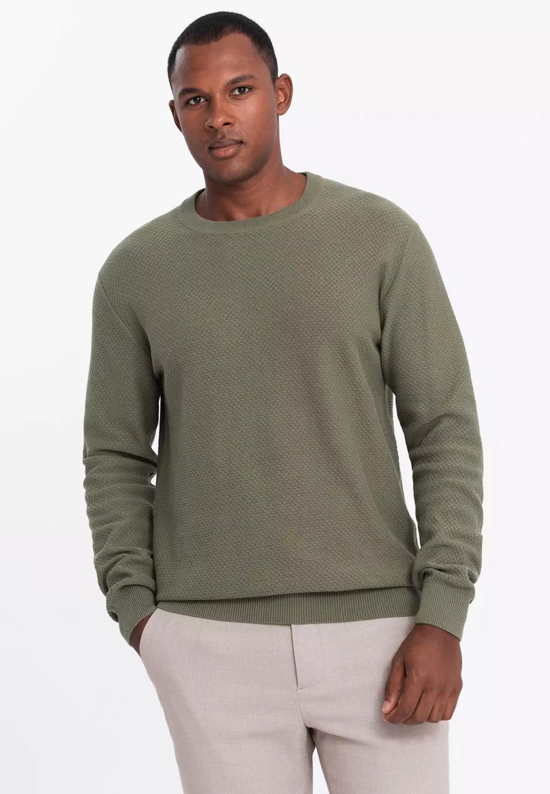 Maglione verde texture con maniche lunghe e scollo rotondo, abbinato a pantaloni chiari. Motivo a maglia grossa su tutto il capo.