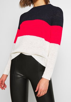 Femme portant un pull en maille colorblock bleu marine, rouge et blanc, associé à un pantalon en cuir noir taille haute sur un fond uni.