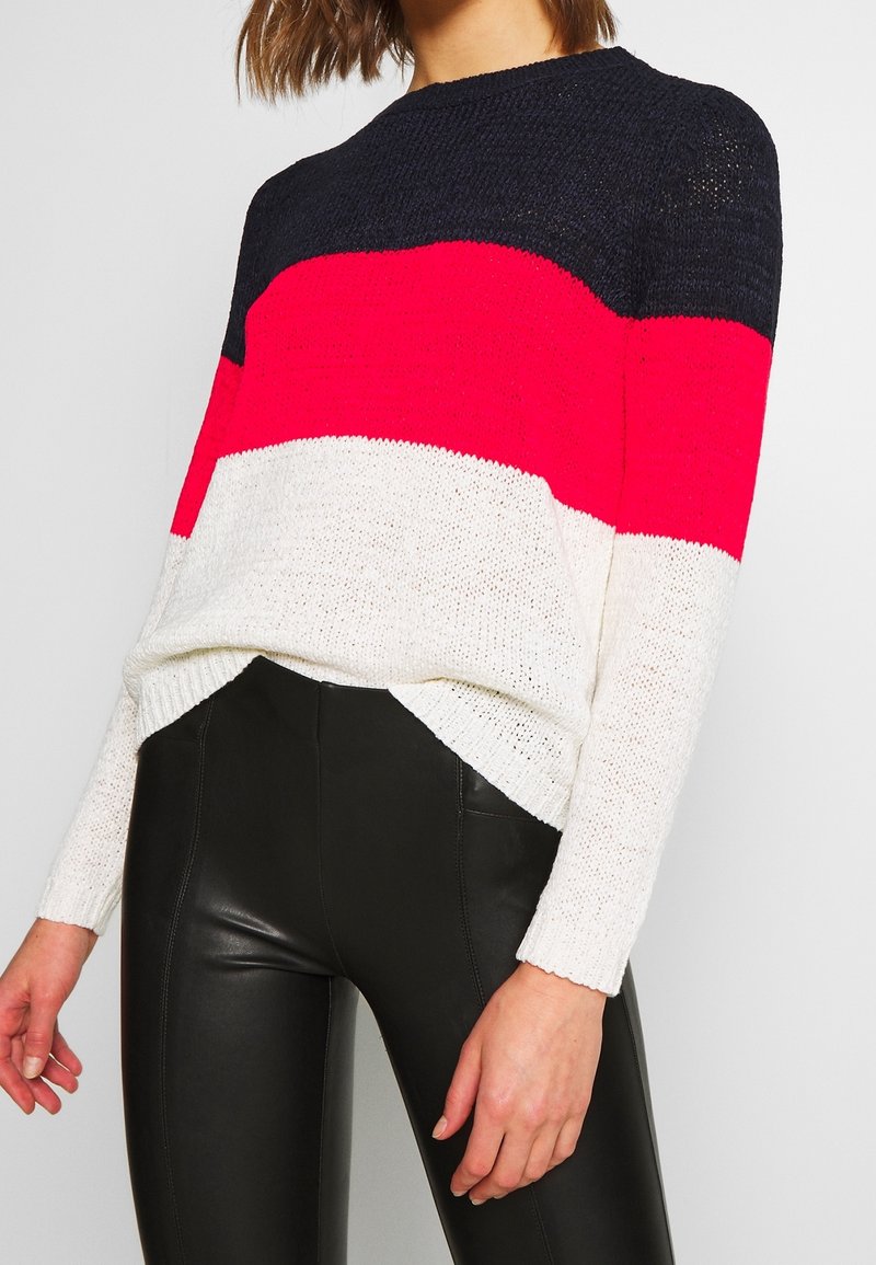 Femme portant un pull en maille colorblock bleu marine, rouge et blanc, associé à un pantalon en cuir noir taille haute sur un fond uni.