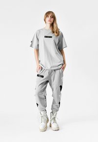 Camiseta gris con un parche de logo negro, combinada con pantalones jogger grises que tienen acentos negros. Botas blancas con cordones completan el conjunto.