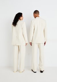 Costume de culoare crem cu sacouri structurate și pantaloni cu croială largă. Sacourile au umeri întăriți și detalii cu nasturi pe manșete.