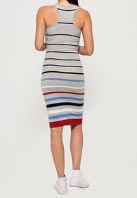 Nainen, joka on pukeutunut hihattomaan, polvipituiseen harmaaseen mekkoon, jossa on vaakasuoria mustia, sinisiä, valkoisia ja punaisia raitoja, ja hänellä on valkoiset tennarit, kuvattuna takaapäin.