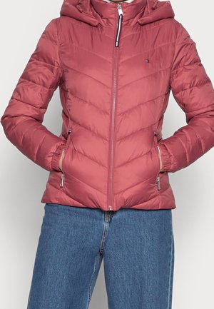 Personne portant une veste matelassée à capuche rose poussiéreuse avec des poches zippées et une fermeture éclair complète à l'avant, associée à un jean en denim bleu.