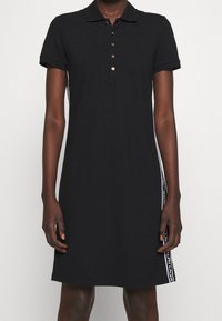 Svart polo-klänning i bomull, med korta ärmar, krage, fem guldknappar och vita sidostreck med textaccenter.