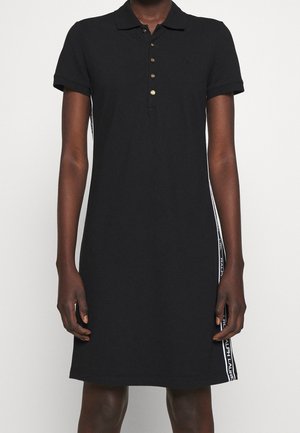 Svart polo-klänning i bomull, med korta ärmar, krage, fem guldknappar och vita sidostreck med textaccenter.